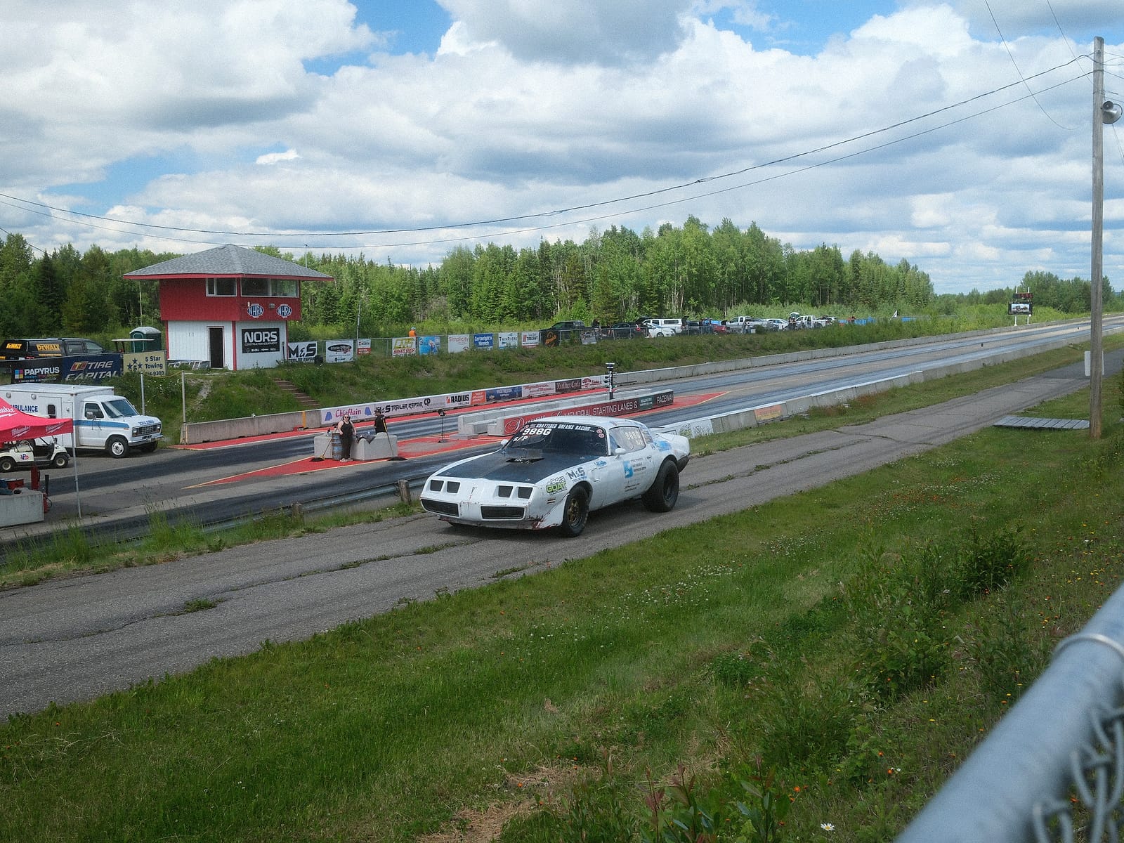 A white door car on the drag strip passing the red timing tower and sponsor boards at NITRO Motorsports Park.